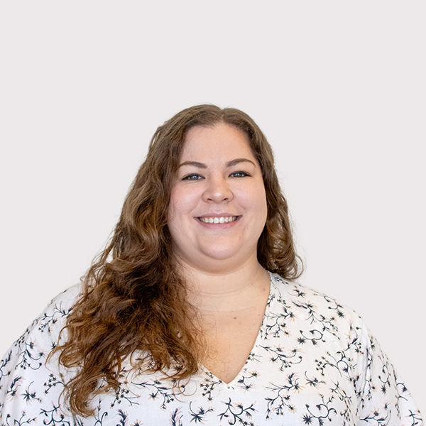 A smiling woman with long curly hair, wearing a floral-patterned blouse, against a plain light background.